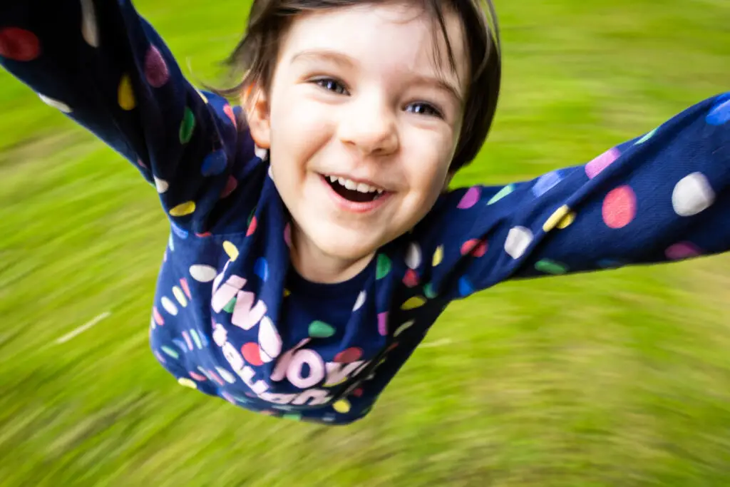 A joyful young child with short brown hair and a colorful polka-dot sweater smiles widely while being twirled around outdoors, with green grass blurred in the background.