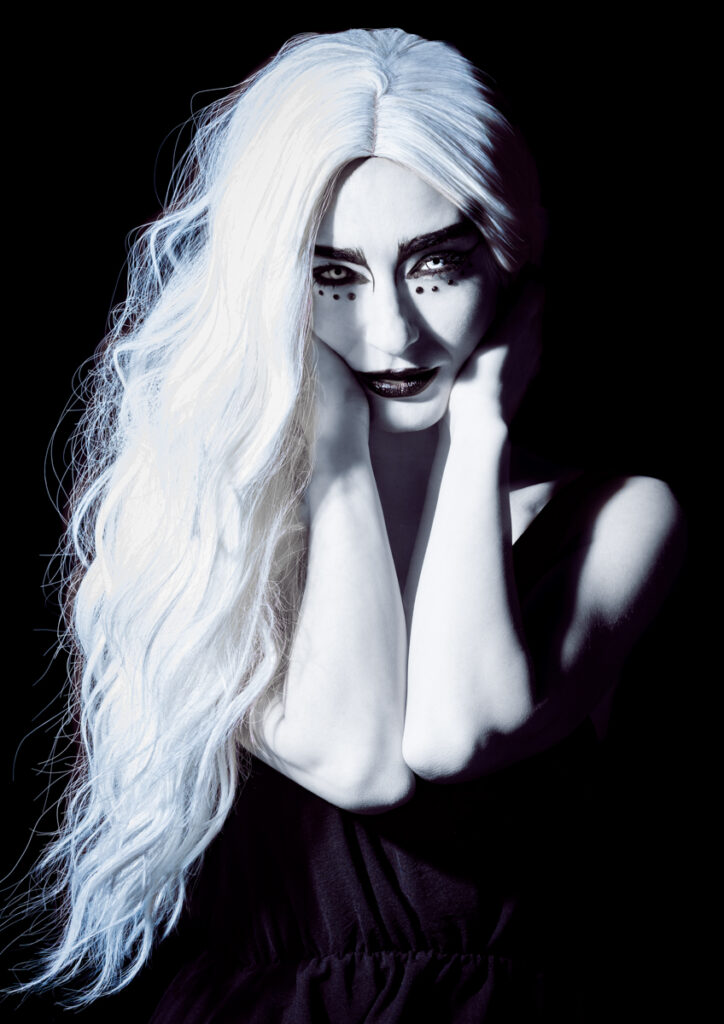 Woman with long white hair and dramatic black-and-white makeup posing in a dark, high-contrast setting.