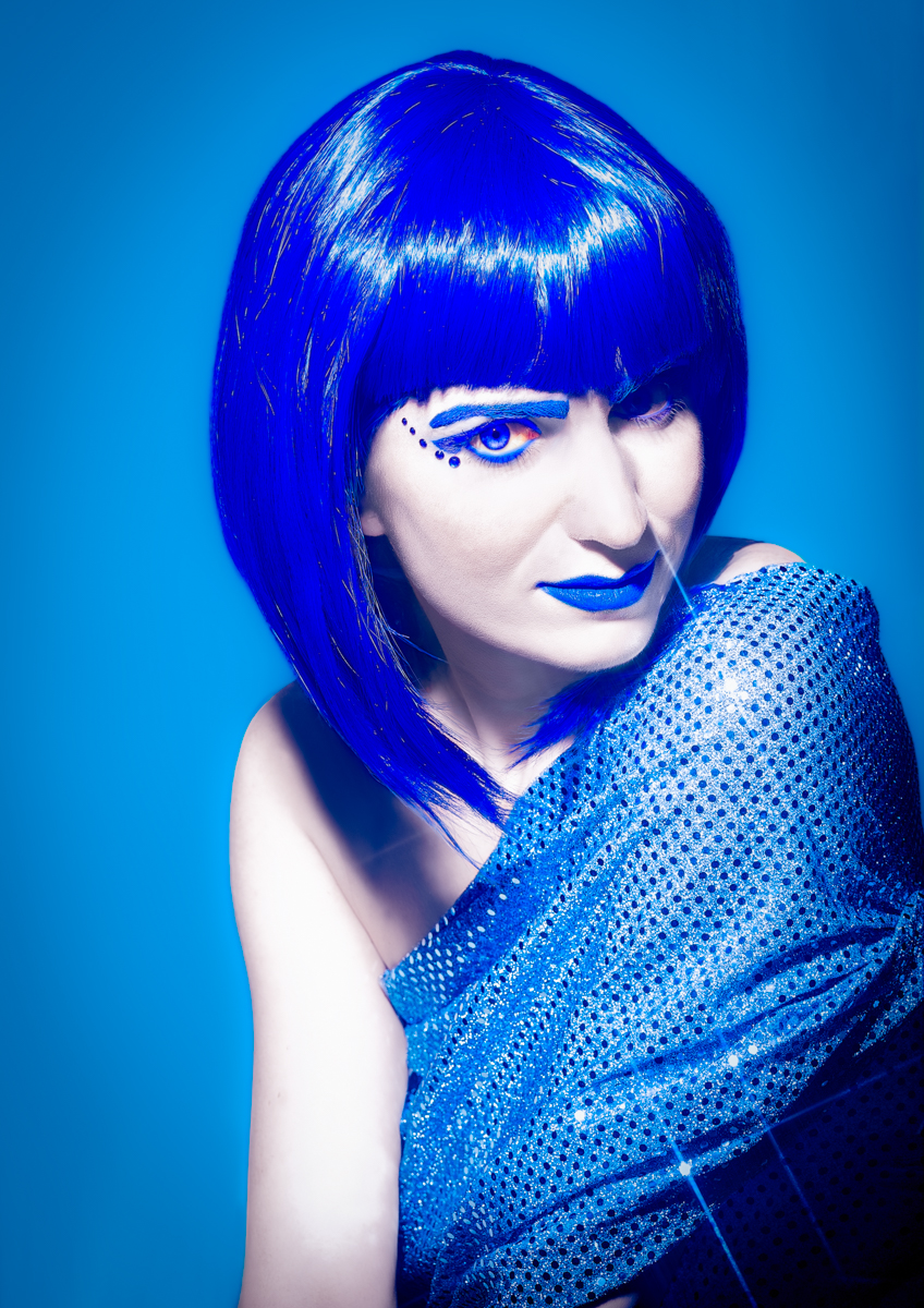 Person with striking blue hair and makeup, wearing a sparkling blue outfit, posing against a vibrant blue background.
