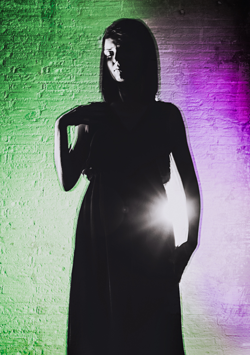 A silhouette of a woman standing in a black dress against a brick wall, illuminated by dramatic green and purple lighting.