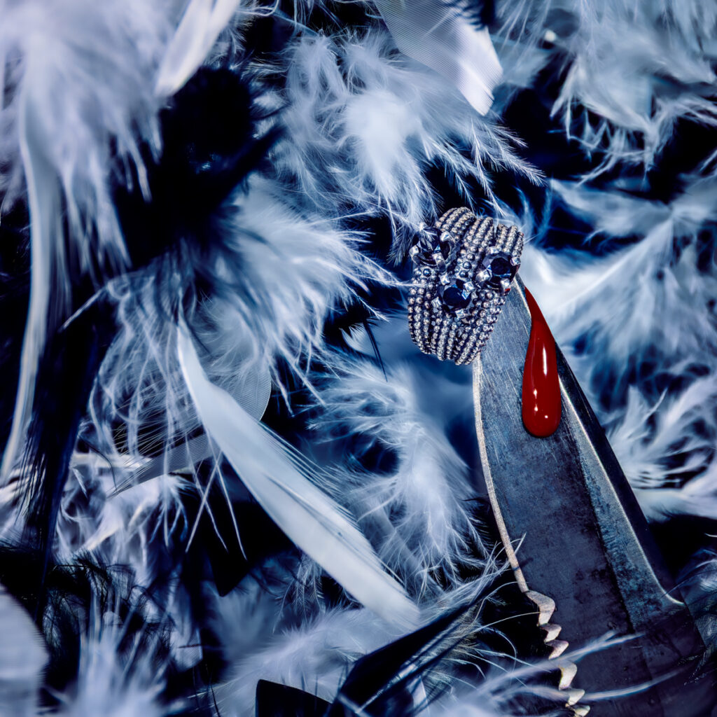 Close-up of a diamond-encrusted ring with blue gemstones held by metallic pliers, a drop of red liquid on the pliers, surrounded by soft white and black feathers.