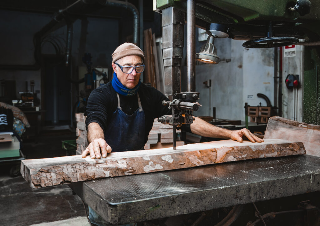 A craftsman wearing safety glasses, a hat, and an apron uses a large saw to cut a thick piece of raw wood inside a woodworking workshop.