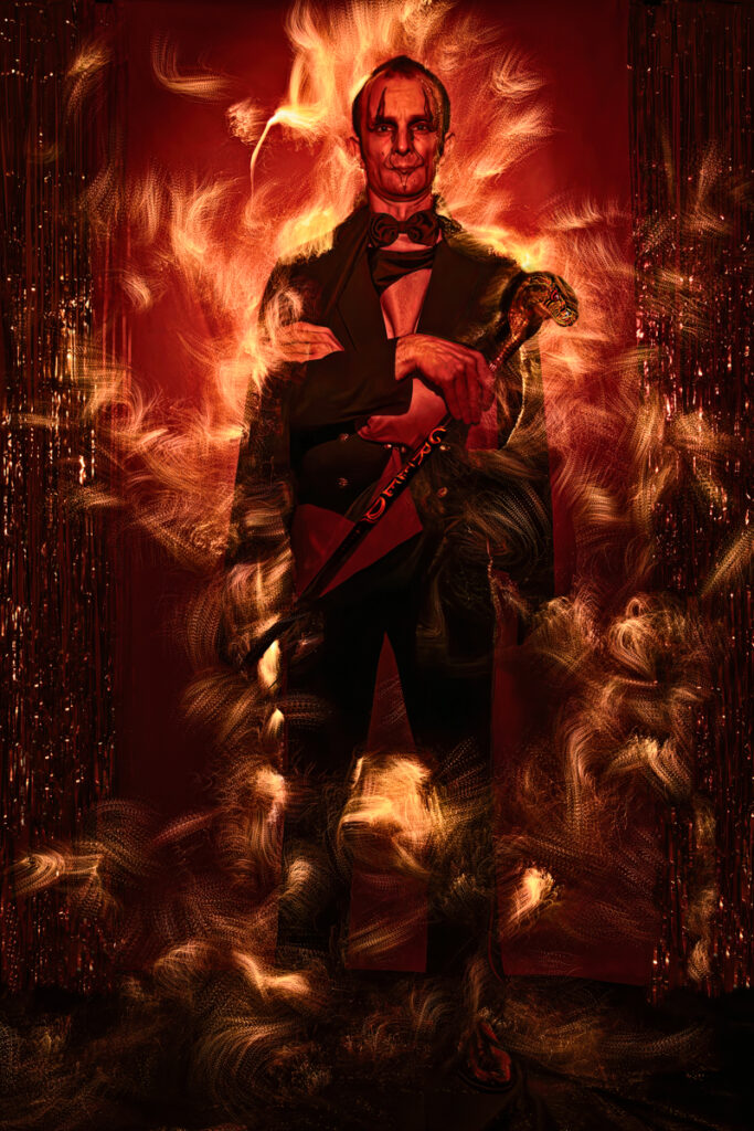 A man in a black suit and tie, partially engulfed in fiery swirls of color, set against a dark red background.