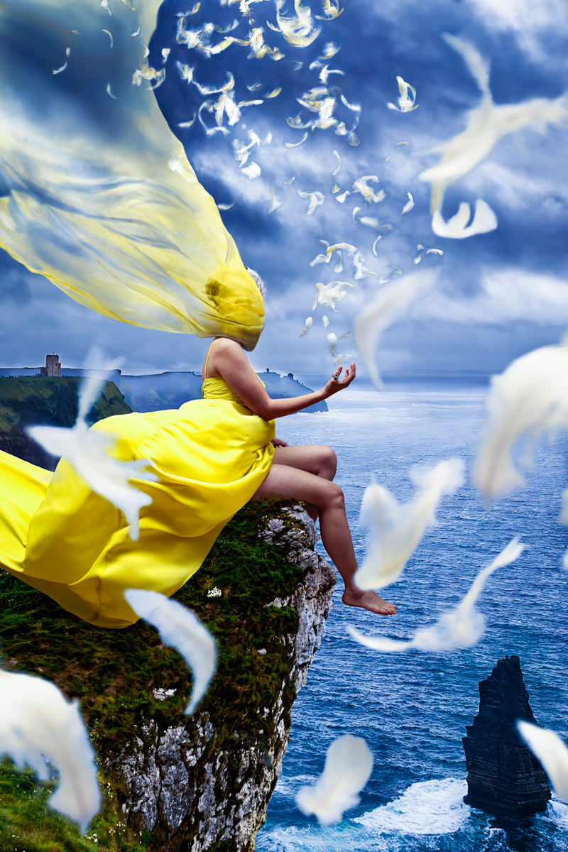A figure in a flowing yellow dress perched on a cliff with billowing fabric and feathers.