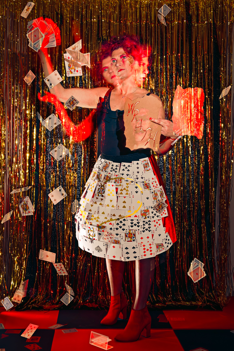 A woman in a black tank top and red shoes stands surrounded by falling playing cards, embodying the Queen of Hearts from Alice in Wonderland, representing the sin of wrath.