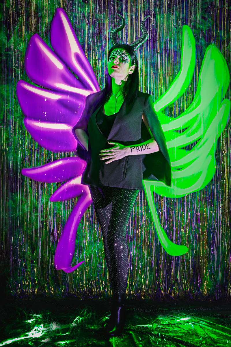 A woman dressed in a black jacket and pants with large wings, standing against a dark green background, representing Maleficent, and embodying the sin of pride.